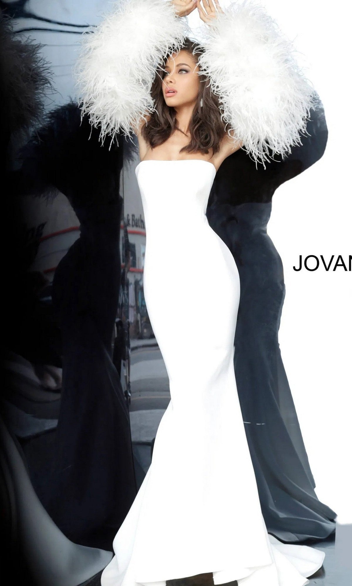 A woman poses in the Jovani 1226 long prom dress, a strapless, form-fitting white gown with dramatic feather sleeves, standing against a white background and partial reflection with